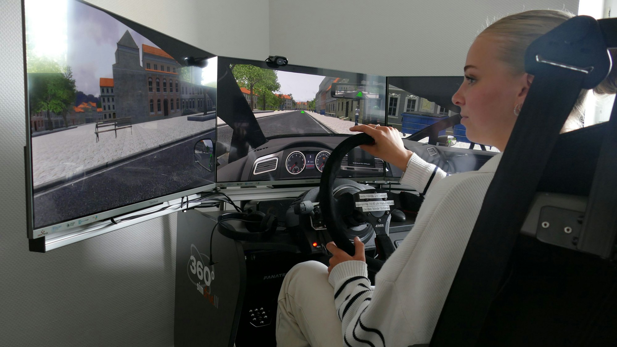Eine junge Frau in weißer Hose und weißem Pullover sitzt auf einem Autositz vor drei Bildschirmen. Sie zeigen eine Straße und das Gelände links und rechts der Straße.
