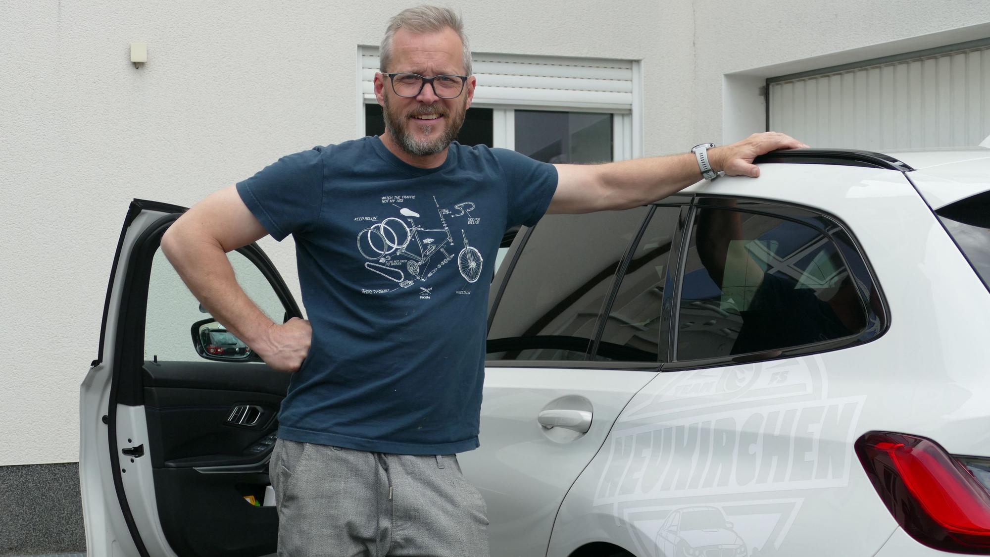 Ein Mann im blauen T-Shirt steht in der geöffneten Tür neben einem weißen Kombi.