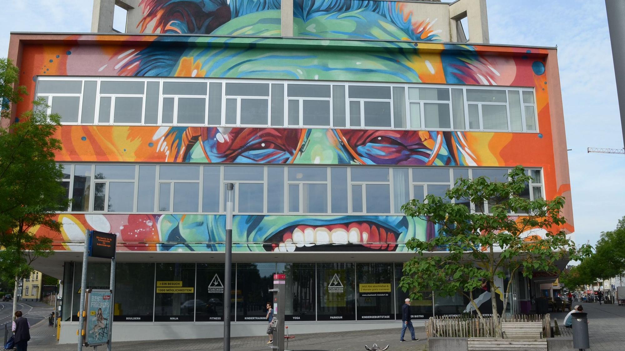 Ein Kaufhausgebäude aus den 70er Jahren. Es ist in grellen Farben bemalt; zu erkennen sind Augen, Zähne und Haare.
