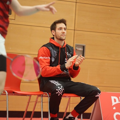 Blick auf einen Sportler, der auf einem Stuhl ein Badmintonspiel verfolgt.