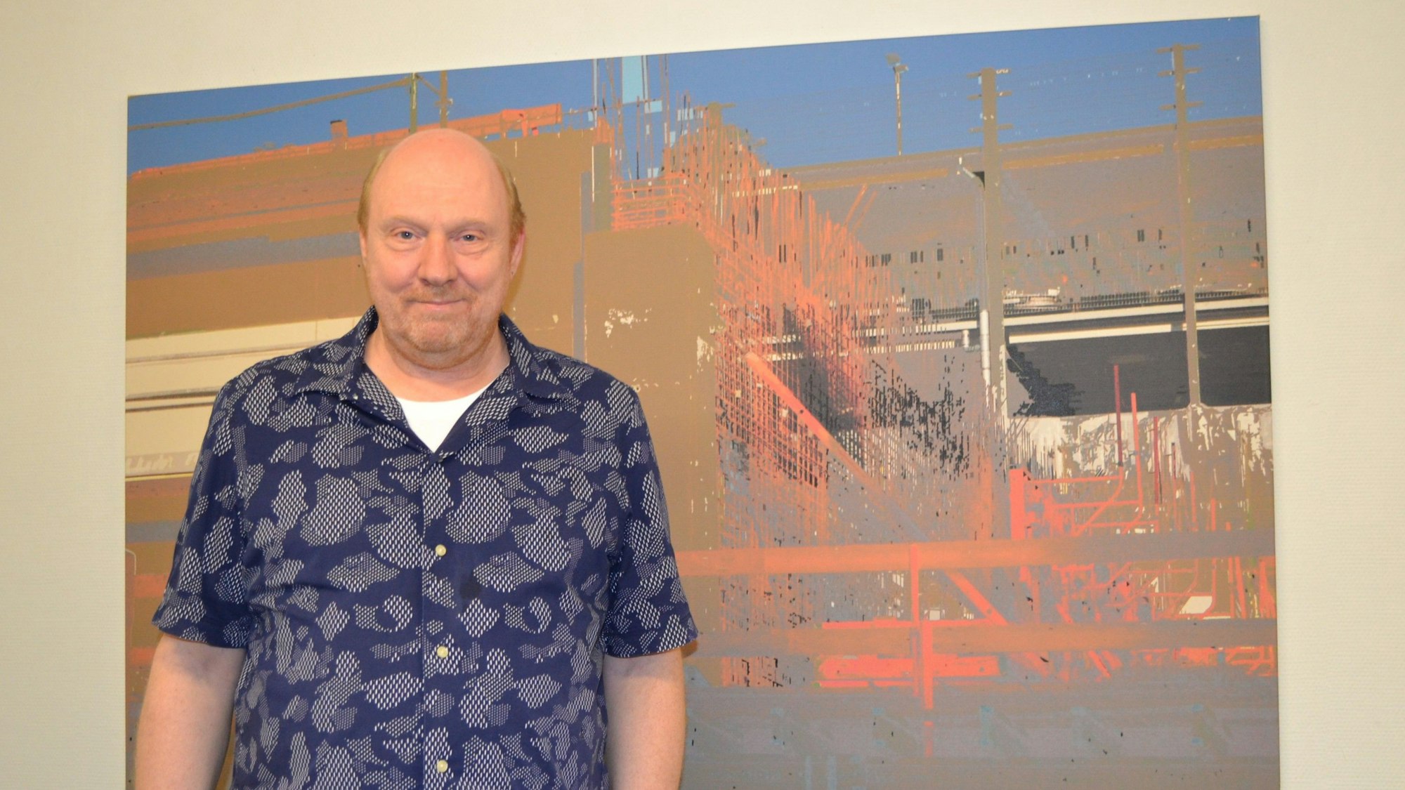Ein Mann im Kurzarmhemd vor einem Kunstwerk an der Wand.
