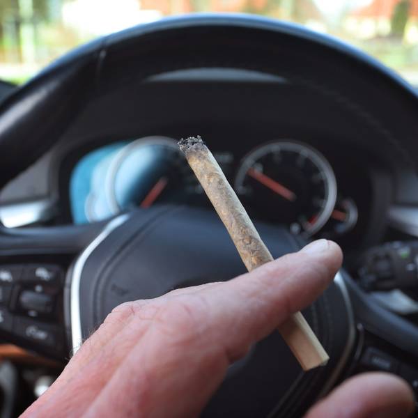 Ein Mann sitzt mit einem Joint zwischen den Fingern am Steuer eines Autos.