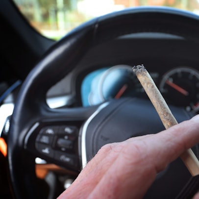 Ein Mann sitzt mit einem Joint zwischen den Fingern am Steuer eines Autos.