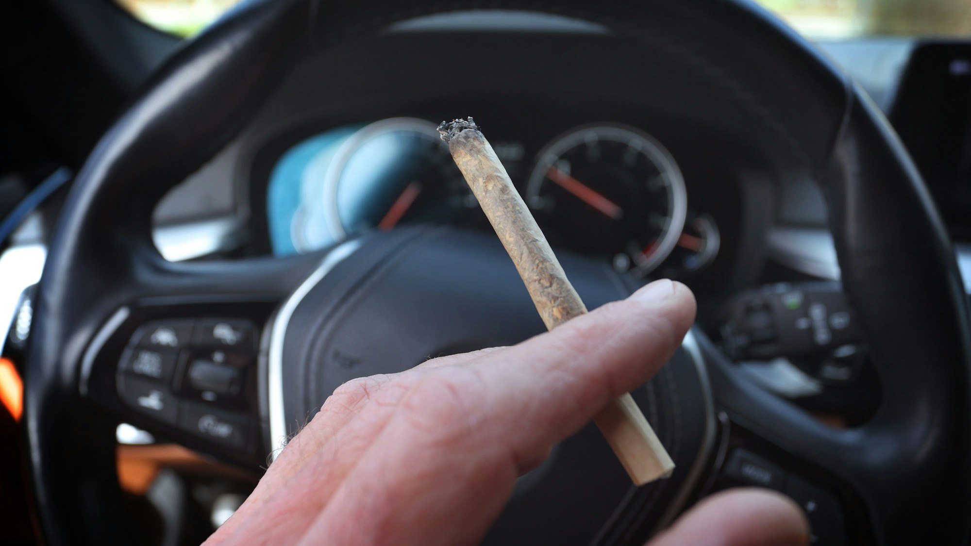 Ein Mann sitzt mit einem Joint zwischen den Fingern am Steuer eines Autos.
