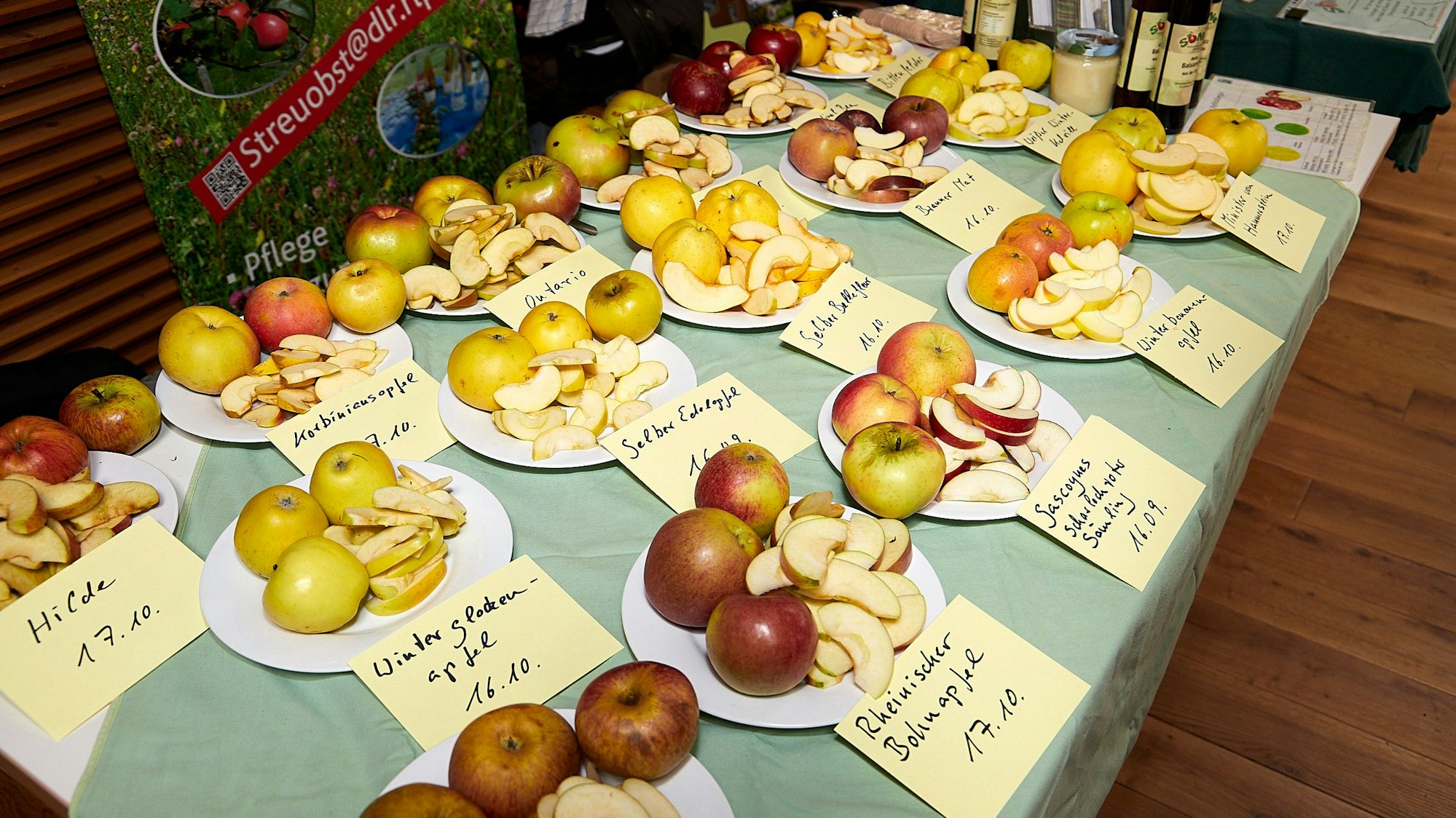 Auf einem Tisch stehen zahlreiche Teller mit Äpfeln. Auf Schildern davor steht jeweils, um welche Sorte es sich handelt.