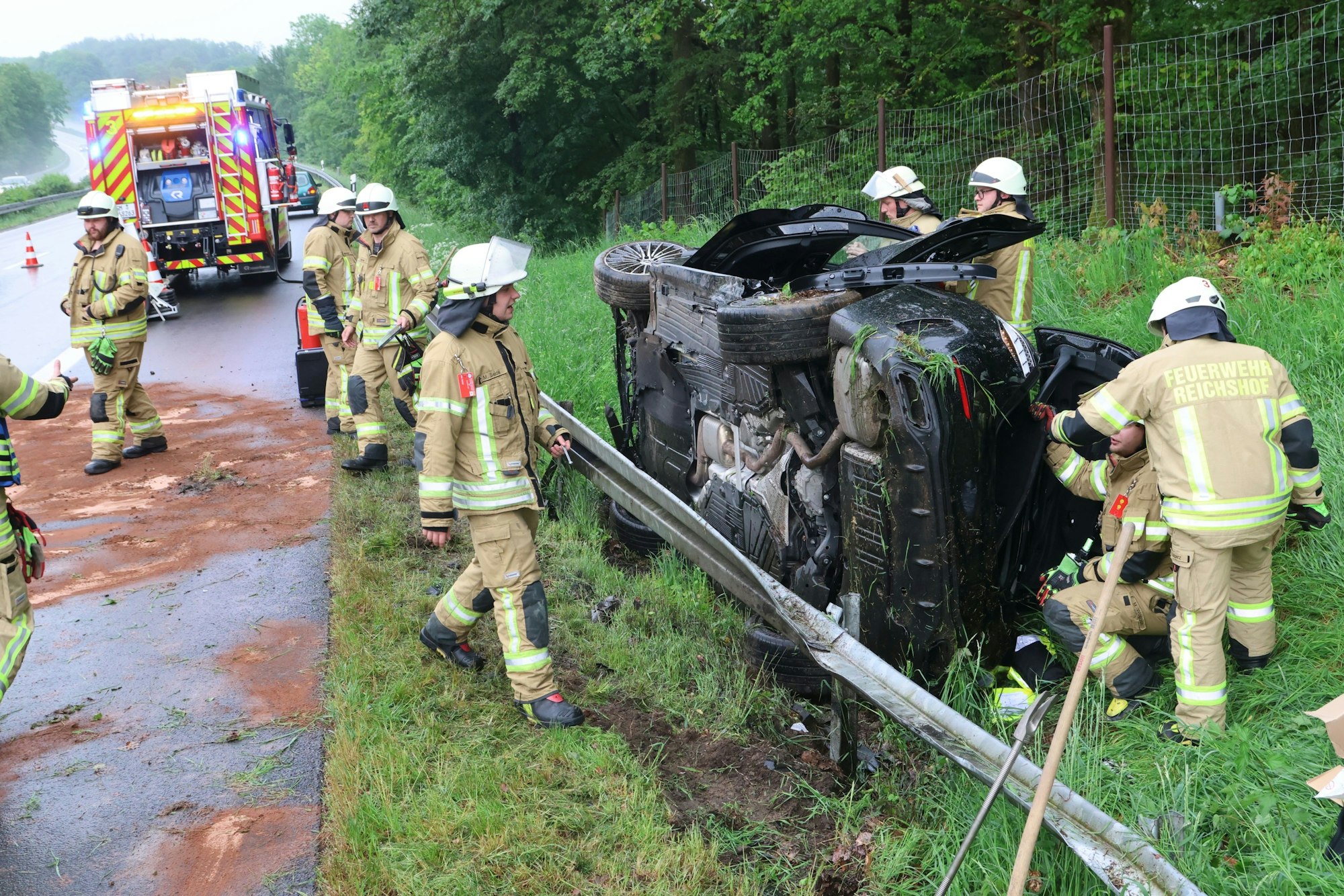 Feuerwehrleute stehen an einem Auto, das hinter der Leitplanke auf der Seite liegt.
