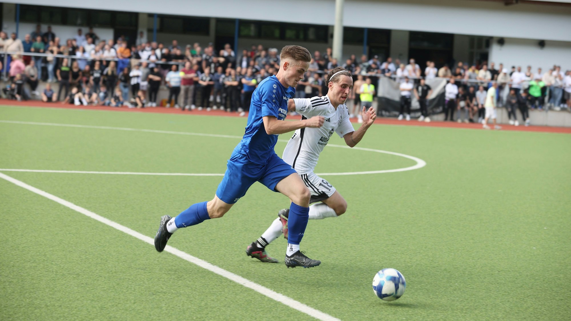Eine Spielsituation zwischen zwei Mannschaften auf einem Fußballplatz. Im Hintergrund sind viele Zuschauer zu sehen.