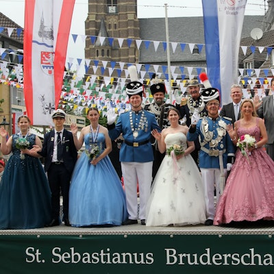 Eine Gruppe von Männern in Schützenuniform und Frauen in Ballkleidern nimmt auf einer Tribüne die Schützenparade ab.