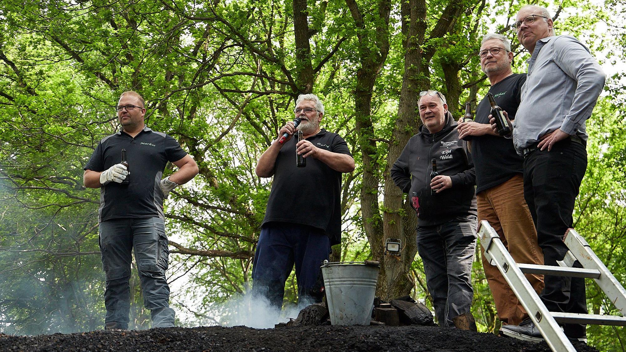 Markus Linden, Gerd Linden, Walter Lehnertz, Michael Lammertz und Jochen Weiler stehen an dem qualmenden Kohlemeiler und halten Bierflaschen in der Hand.
