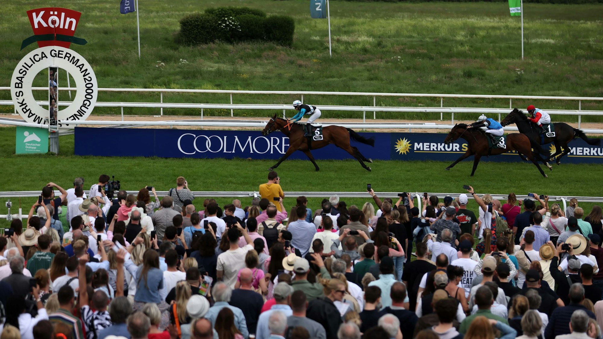 Man sieht sich und wird gesehen: Der traditionelle Pfingst-Ausflug zur Rennbahn hat nichts von seiner Faszination verloren. Drei Pferde im Zieleinlauf, mit größeren Abständen, davor schaut ihnen eine große Menschenmenge zu.