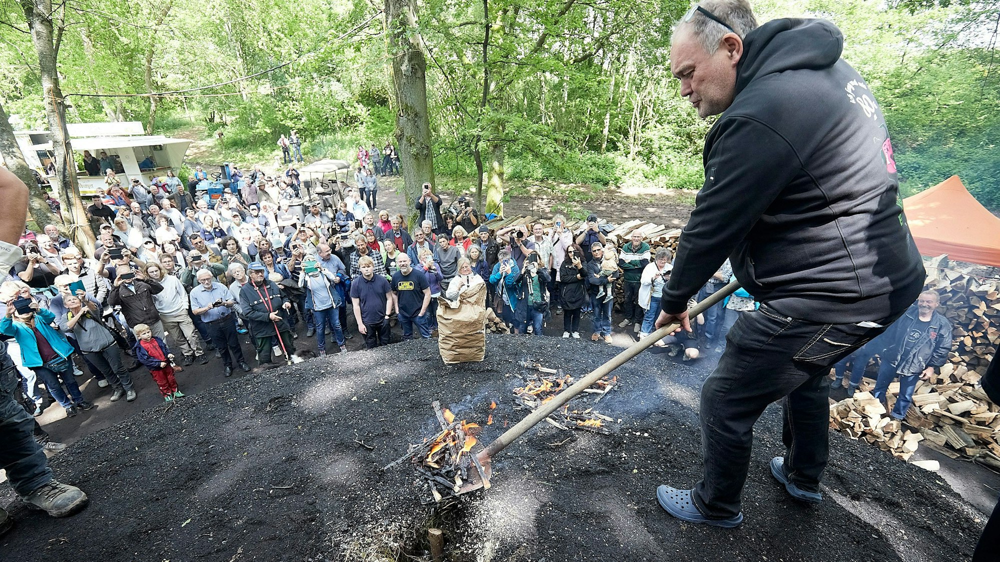 Walter Lehnertz steht auf einem schwarzen Hügel und hält eine Schaufel mit Glut in der Hand. Rund um den Kohlemeiler stehen zahlreiche Zuschauerinnen und Zuschauer.