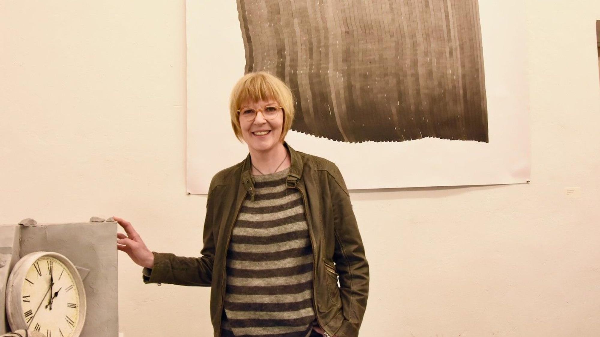 Eine Frau mit Pagenkopf-Frisur steht vor einem Gemälde, das an der Wand hängt und neben einer Wanduhr.