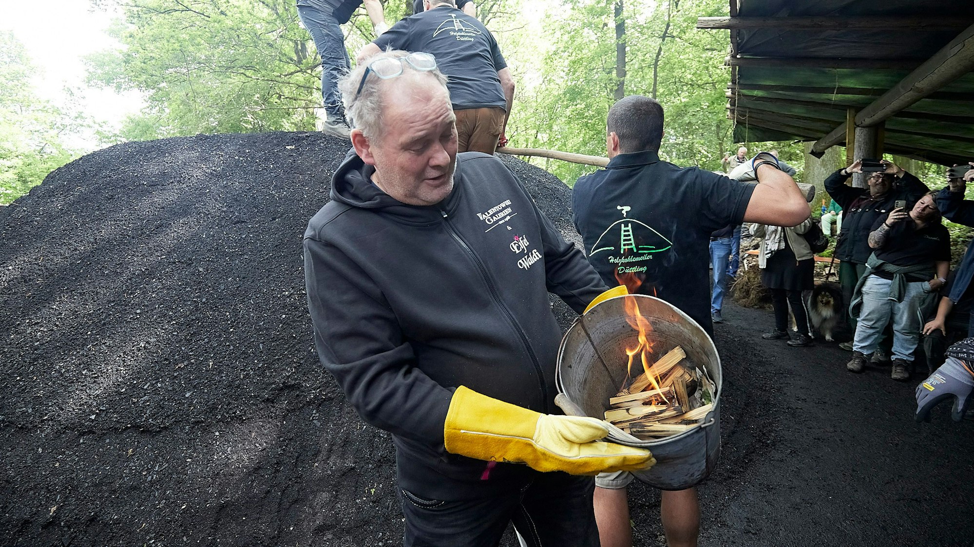 Walter Lehnertz hält einen Heimer mit brennenden Holzscheiten in der Hand. Im Hintergrund ist der Holzkohlemeiler zu sehen.