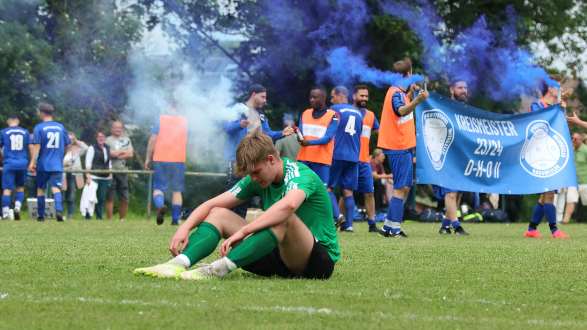 Ein Spieler einer Fußballmannschaft sitzt enttäuscht auf dem Boden, die Spieler der anderen Mannschaft feiern im Hintergrund mit Rauchfackeln.