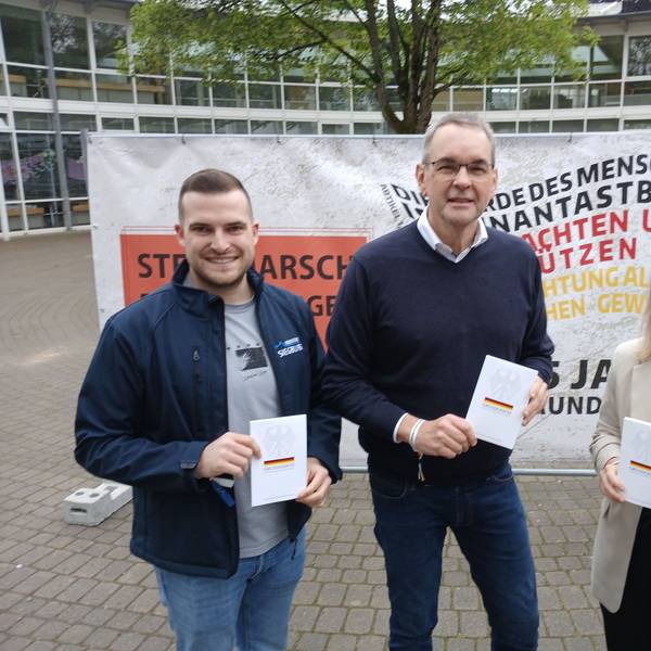 Zwei Männer und eine Frau mit Büchern in der Hand vor einem Plakat