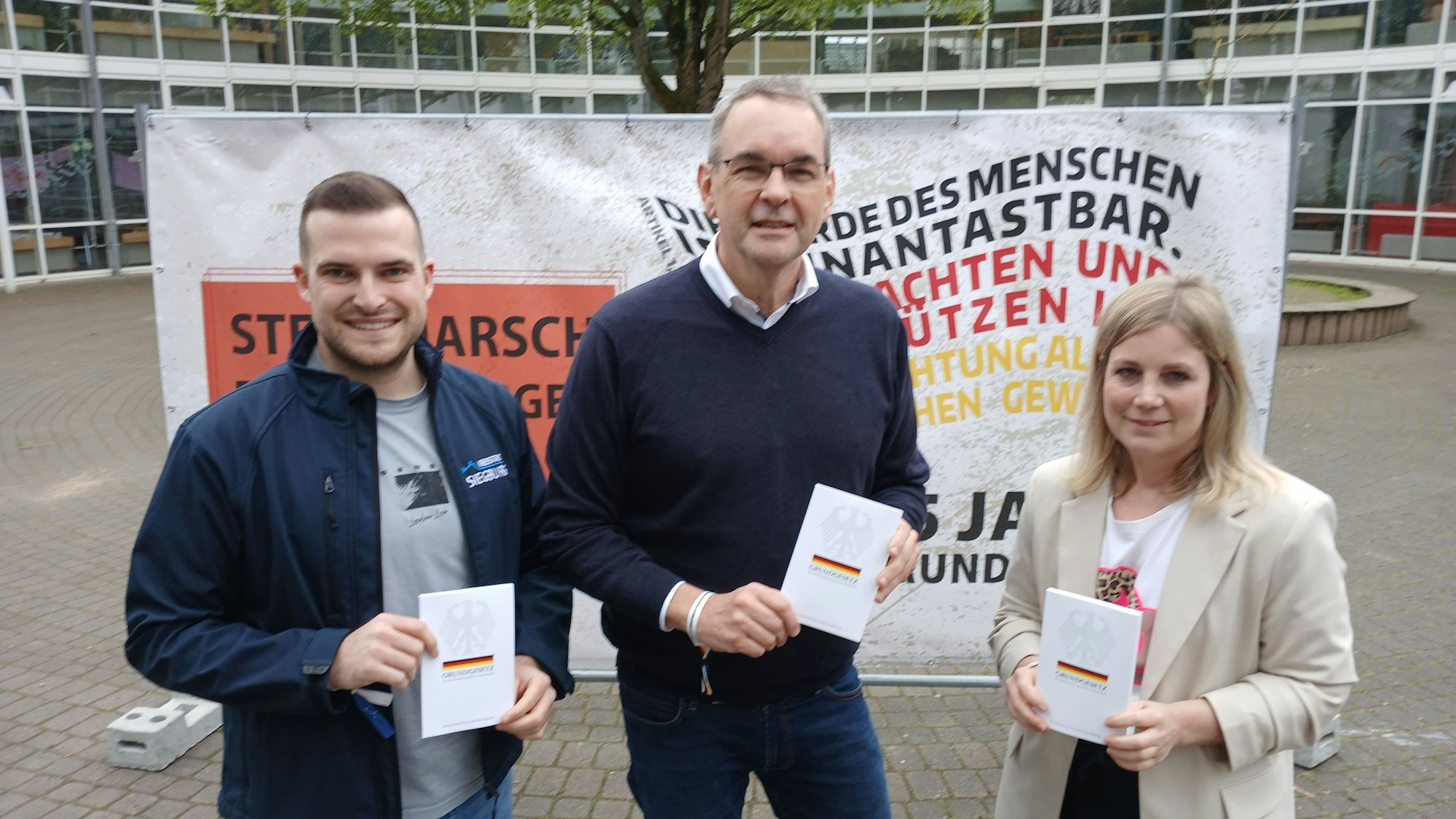 Zwei Männer und eine Frau mit Büchern in der Hand vor einem Plakat