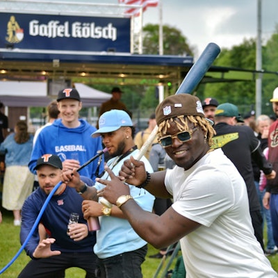 Ein Spieler mit blonden Rastalocken unter einer Kappe hält einen Baseballschläger in den Händen, im Hintergrund zieht jemand am Schlauch einer Bong.