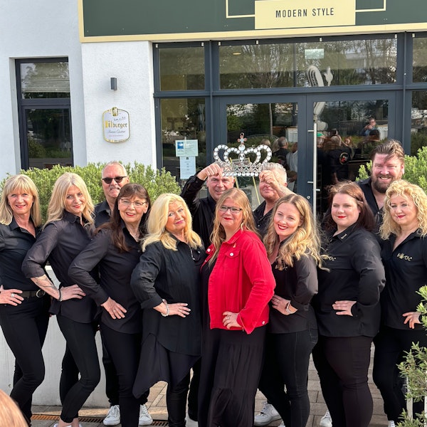 Eine Gruppe von schwarz gewandeten Damen und Herren, in der Mitte die künftige Prinzessin in rotem Blazer und Krone, die von zwei Männern über ihrem Kopf gehalten wird.