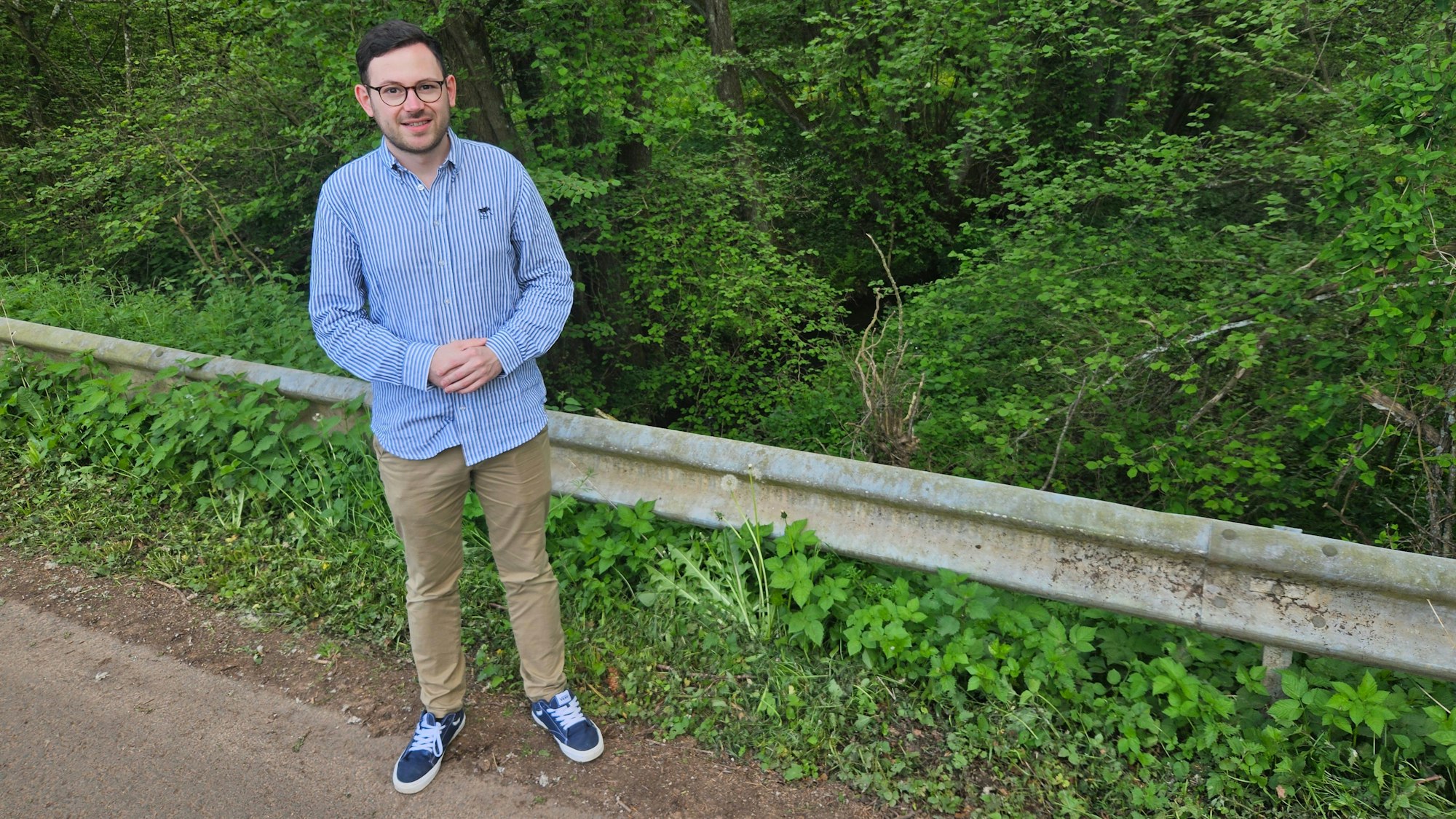Emmanuel Kunz, der Ortsvorsteher von Golbach, steht auf einer Straße an einer Leitplanke. Im Hintergrund sind viele grüne Bäume und Sträucher zu sehen.