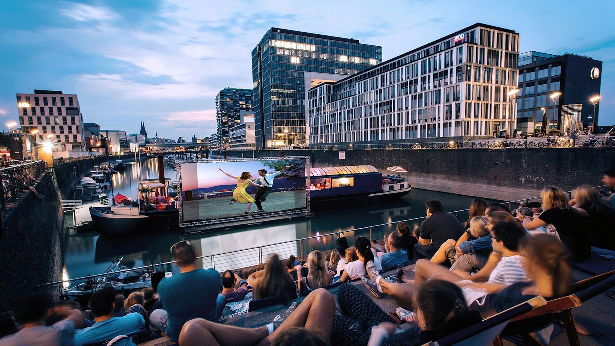 Menschen schauen sich im Open-Air-Kino am Rheinauhafen in Köln vor der Domkulisse einen Film an.