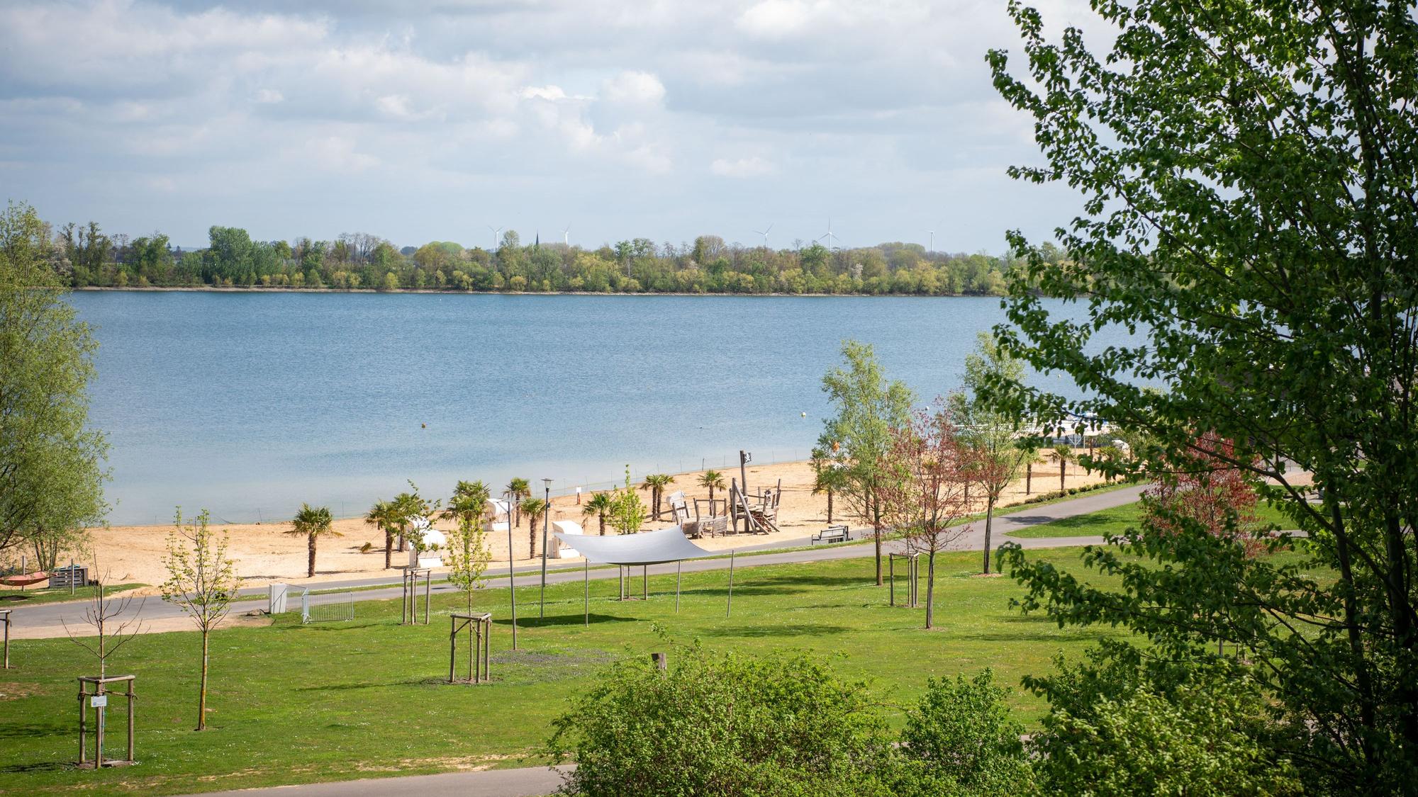 Das Bild zeigt den Wassersportsee in Zülpich mit Sandstrand und Palmen im Frühling.