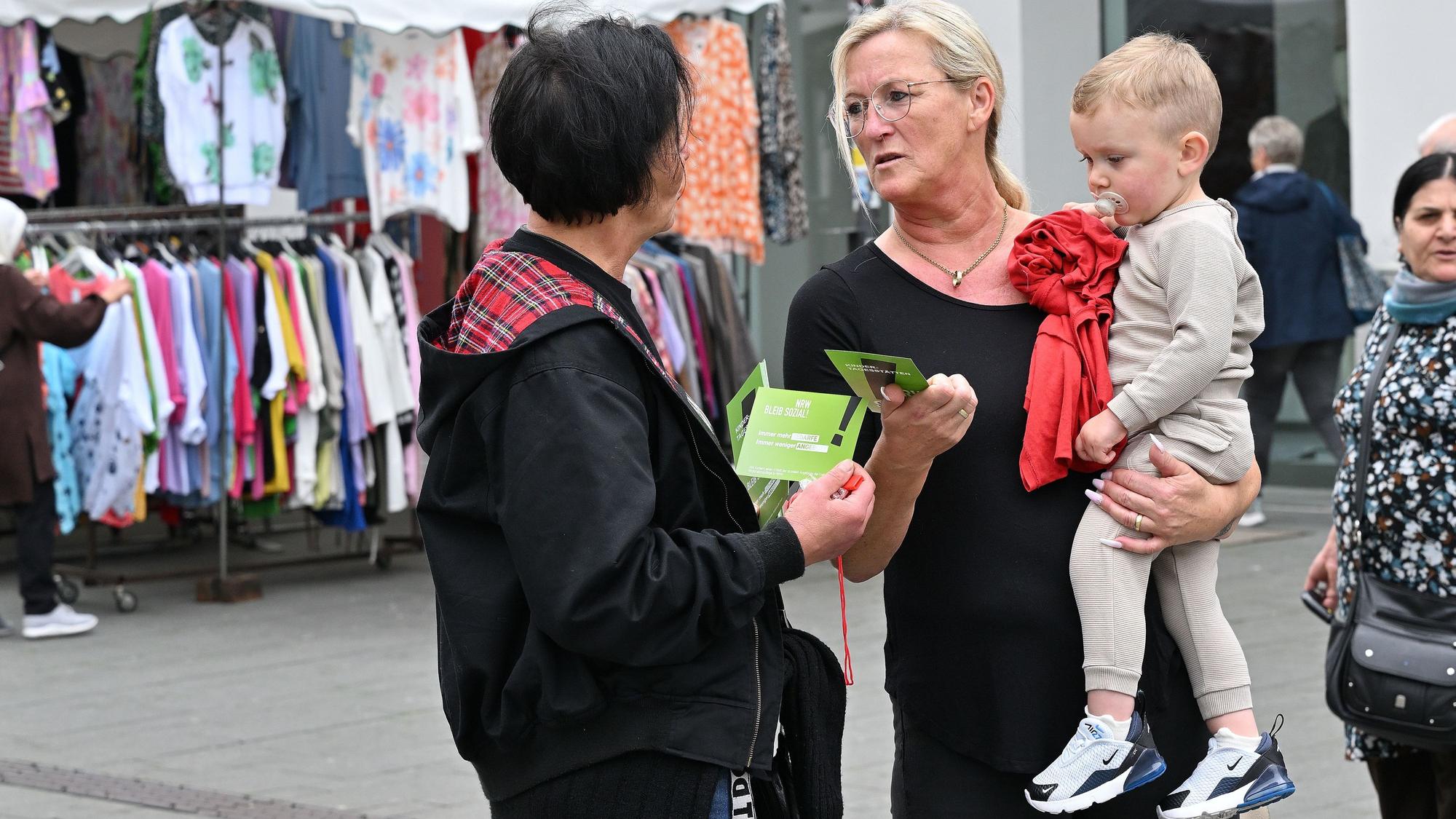 Eine Erzieherin verteilt einen Flyer an eine Frau mit einem Kleinkind auf dem Arm.