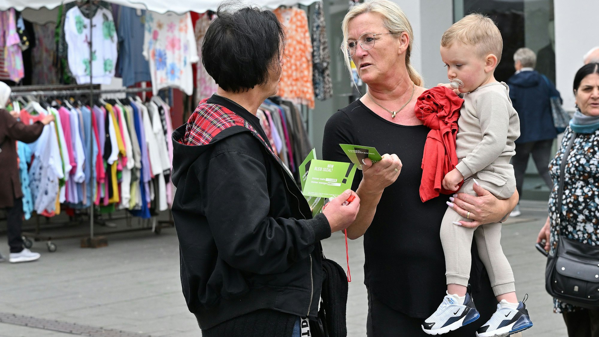 Eine Erzieherin verteilt einen Flyer an eine Frau mit einem Kleinkind auf dem Arm.