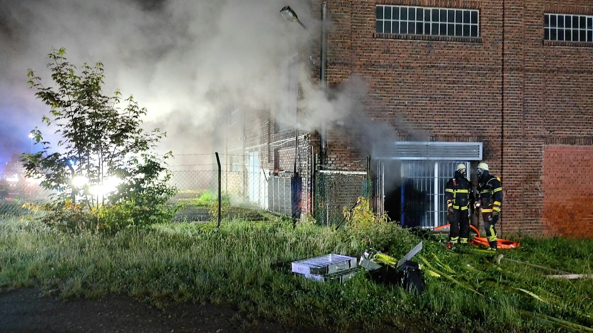 Das Bild zeigt zwei Feuerwehrleute vor der Lagerhalle. Aus den Fenstern dringt Rauch nach außen.