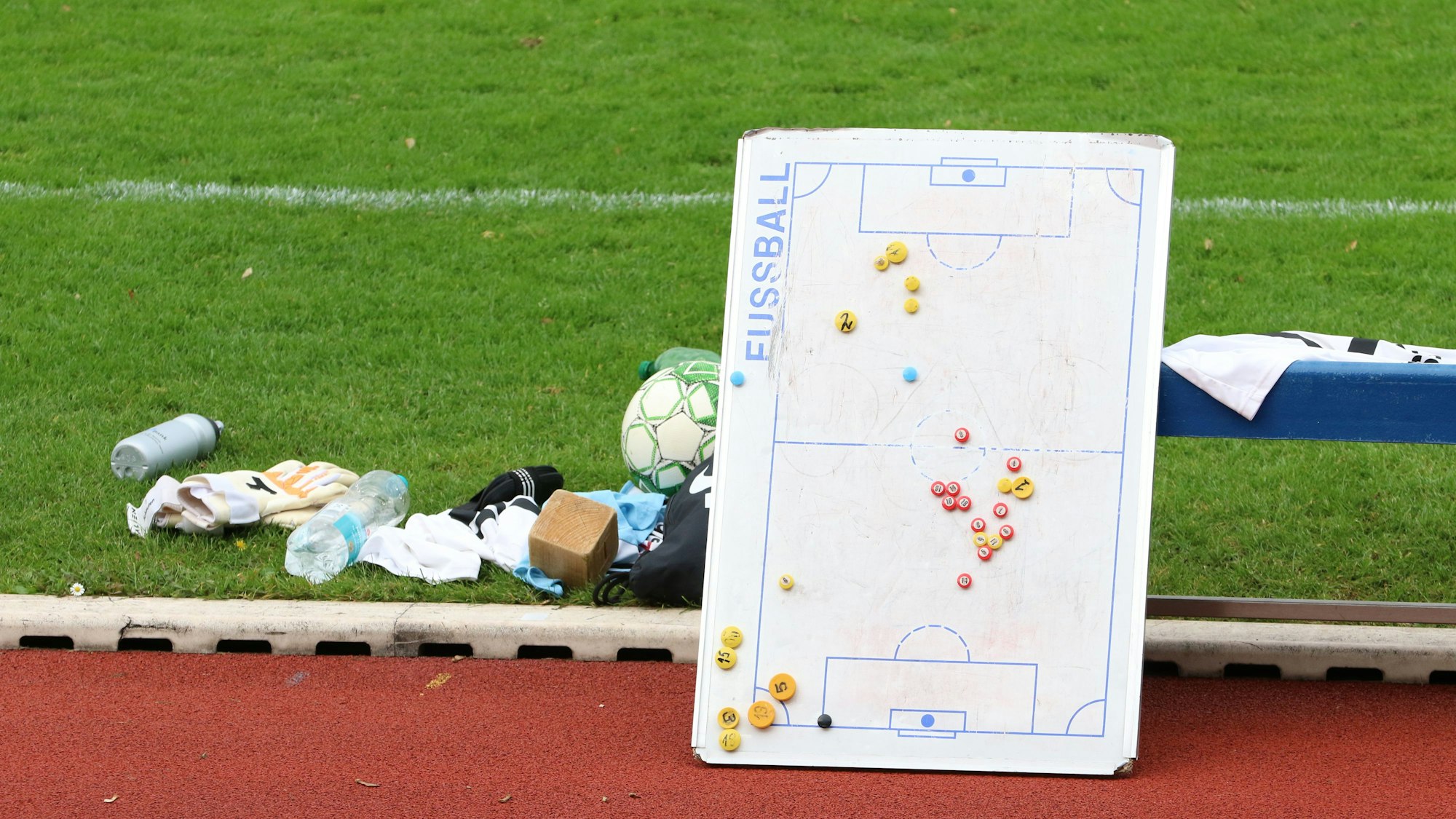 Eine Taktiktafel lehnt an einer Bank, am Rande des Spielfelds liegen Fußballutensilien.
