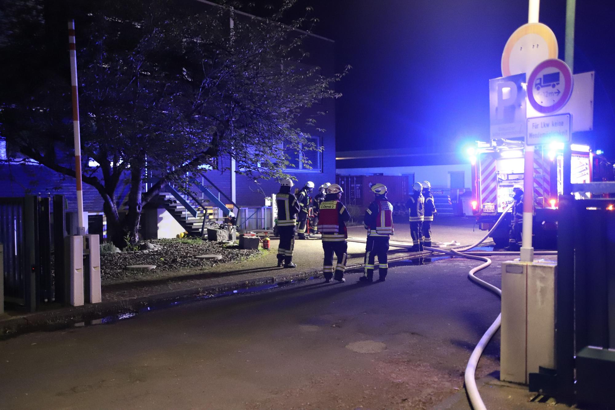 Feuerwehrleute stehen vor den Gebäuden eines Maschinenbauunternehmens und haben Wasserschläuche verlegt.
