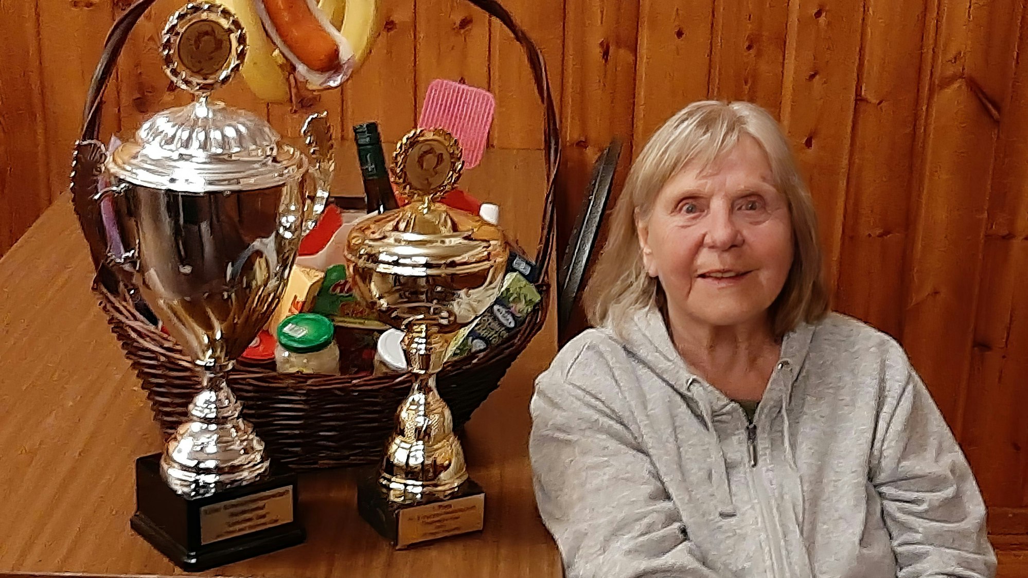 Die 80 Jahre Anita Groll aus Kall sitzt an einem Tisch, auf dem zwei große Pokale und ein Präsentkorb stehen, die sie als Siegerin der Schockermeisterschaft in Kall gewonnen hat.