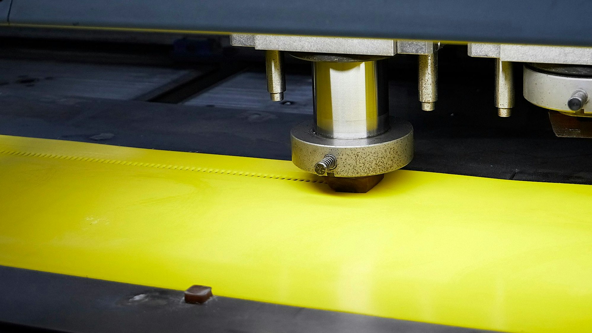Die Detailaufnahme zeigt eine silberne Maschine, die Löcher in ein gelbes Kunststoffband stanzt.