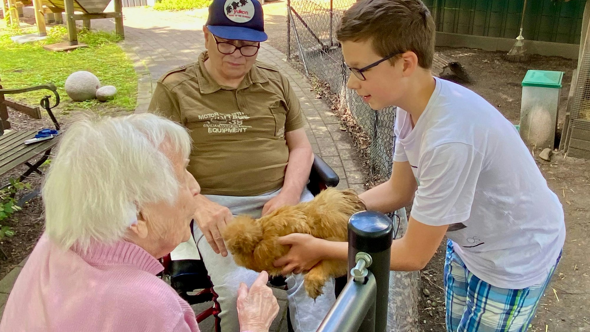 Zu sehen sind zwei Senioren und ein Junge, die eines der Hühner streicheln.