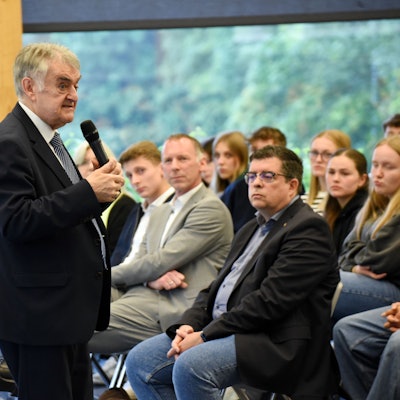 Minister Reul steht mit einem Mikro vor der Schülerschaft. In der ersten Reihe sitzt der Bürgermeister.