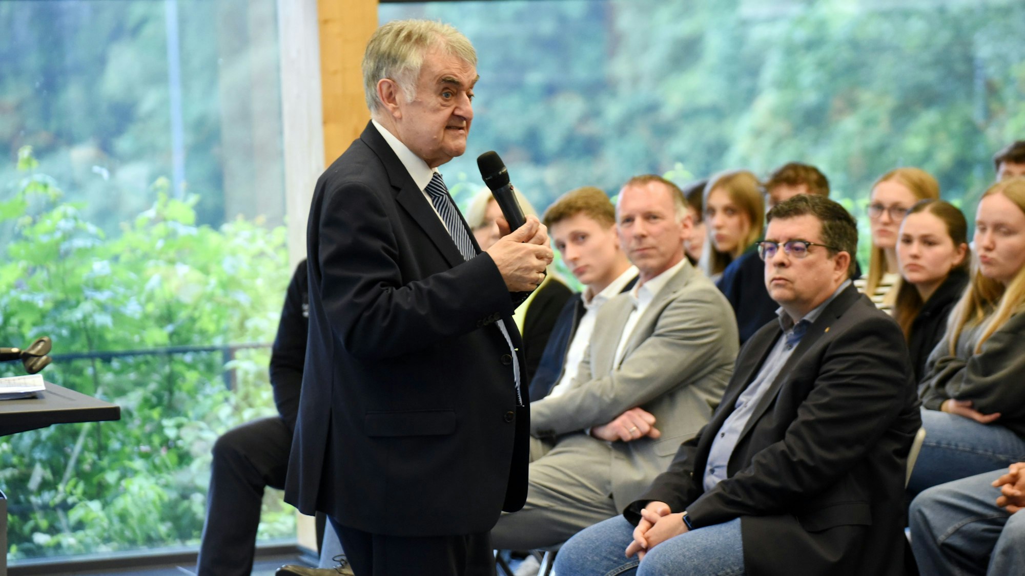 Minister Reul steht mit einem Mikro vor der Schülerschaft. In der ersten Reihe sitzt der Bürgermeister.