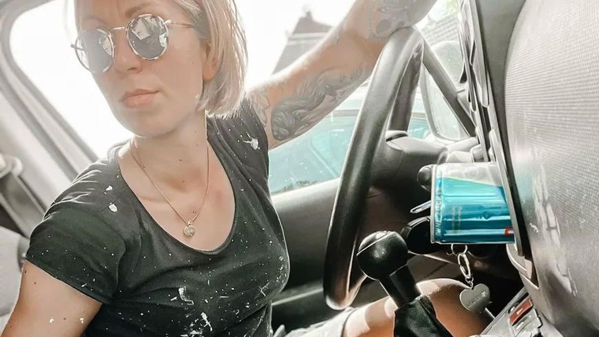 Eine Malerin mit weißen Farbflecken auf Armen und Kleidung sitzt am Steuer eines Autos.
