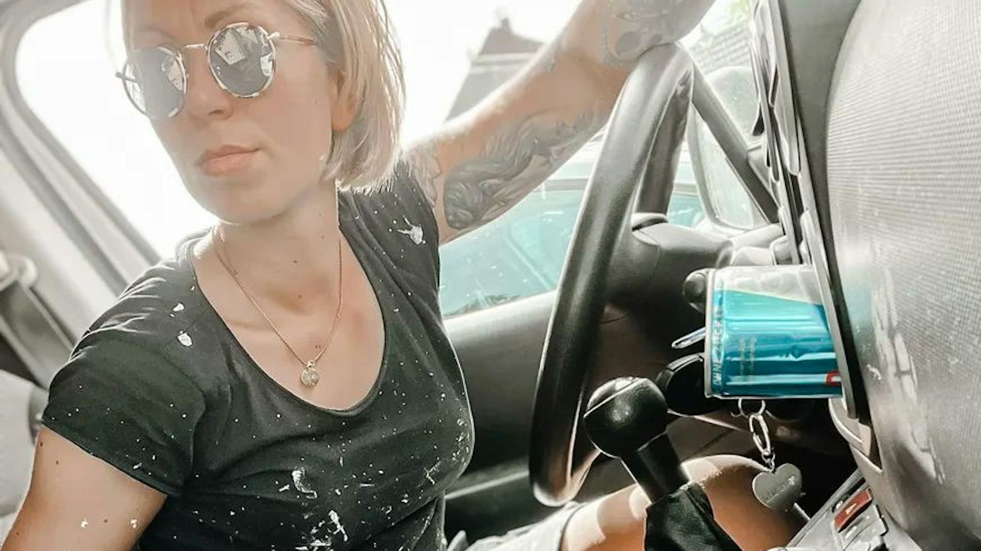 Eine Malerin mit weißen Farbflecken auf Armen und Kleidung sitzt am Steuer eines Autos.