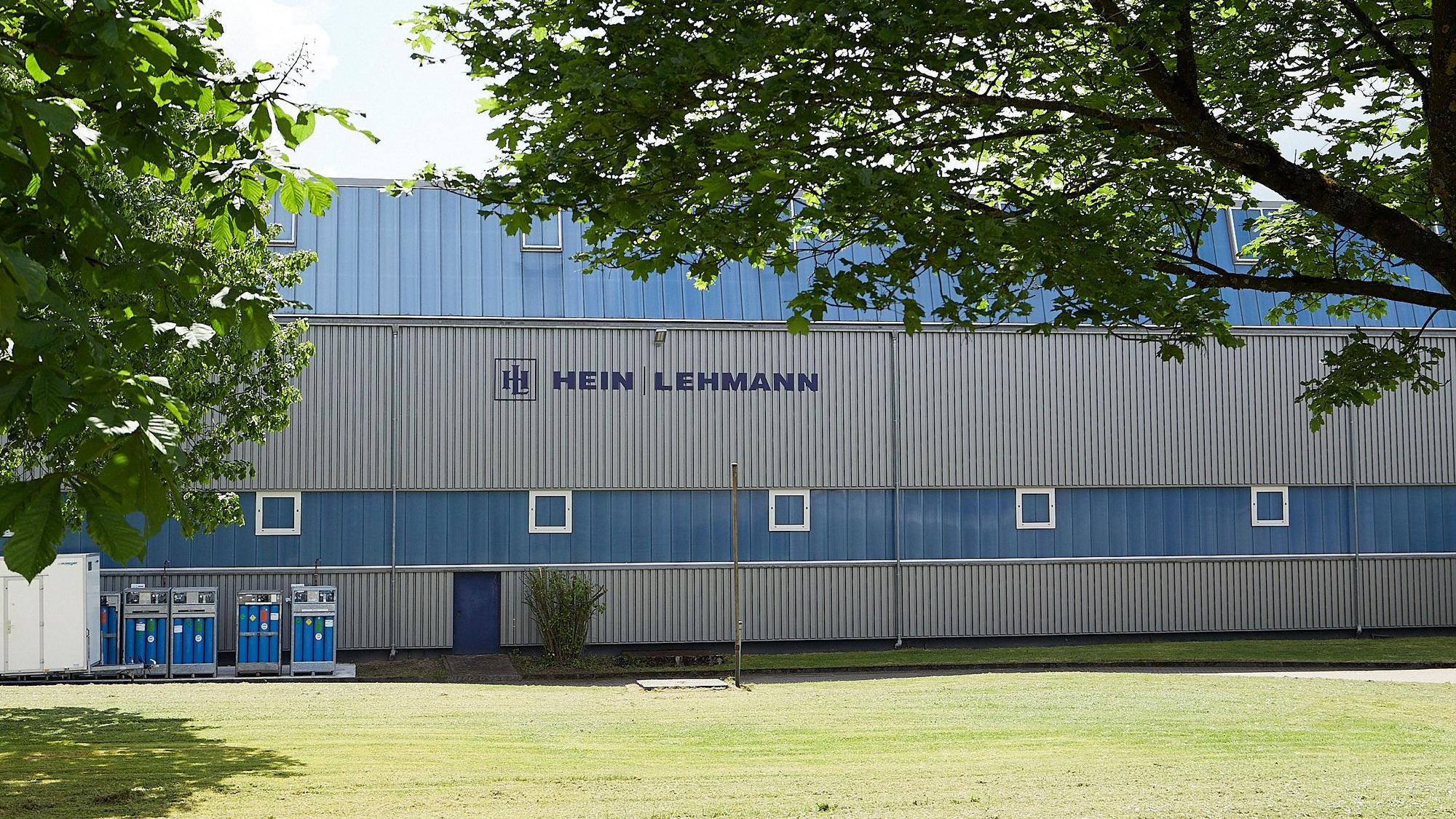 Hinter zwei Bäumen liegt eine der blau-grauen Produktionshallen der Firma Hein, Lehmann in Zingsheim.