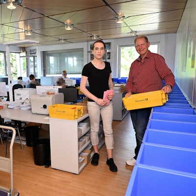 In dem Raum gibt es viele Computer-Arbeitsplätze. Frank Bodengesser hält einen gelben Cotainer mit versandfertigen Briefen in der Hand. Eine Mitarbeiterin zwei Umschläge für die Briefwahl.