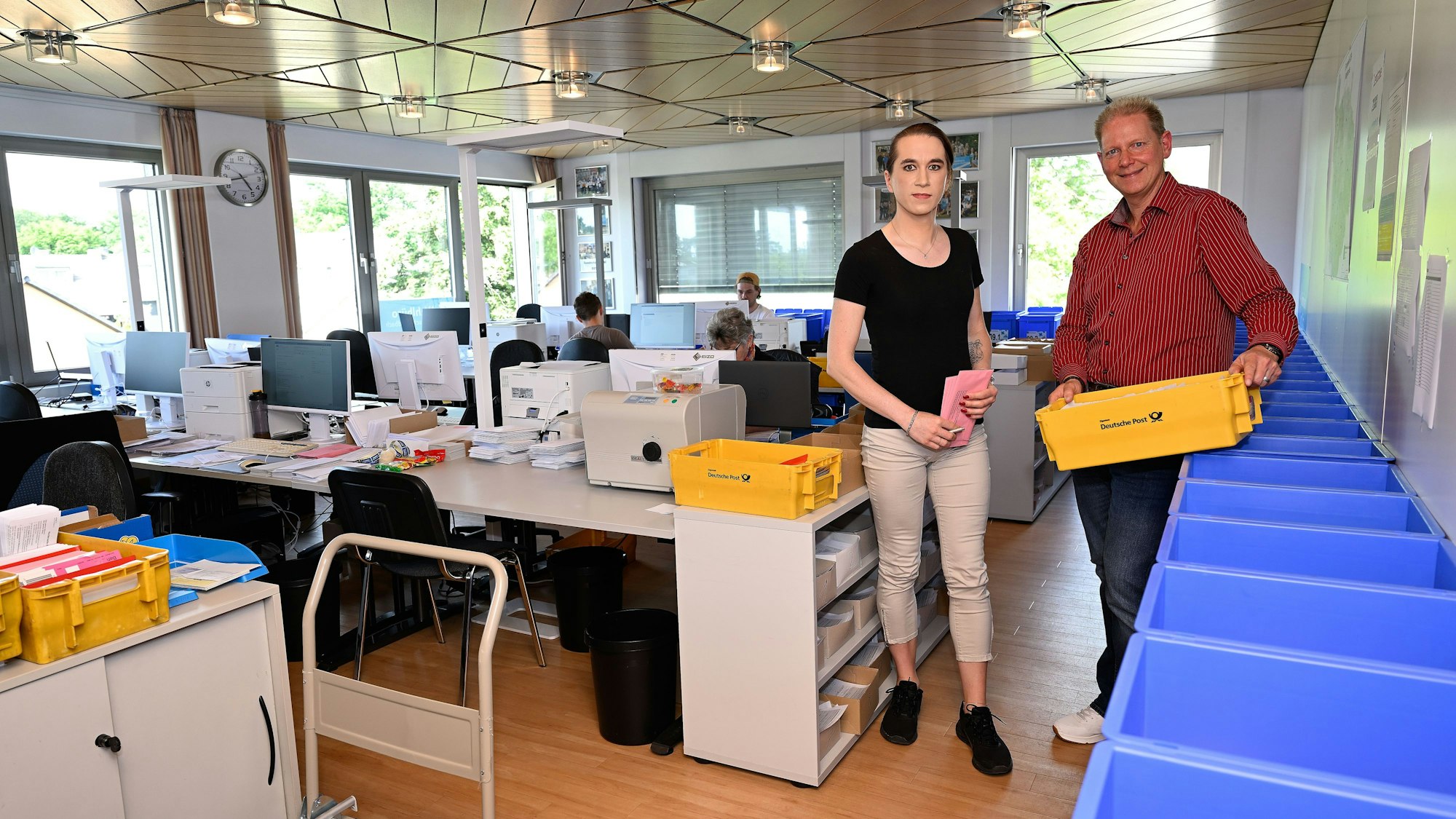 In dem Raum gibt es viele Computer-Arbeitsplätze. Frank Bodengesser hält einen gelben Cotainer mit versandfertigen Briefen in der Hand. Eine Mitarbeiterin zwei Umschläge für die Briefwahl.