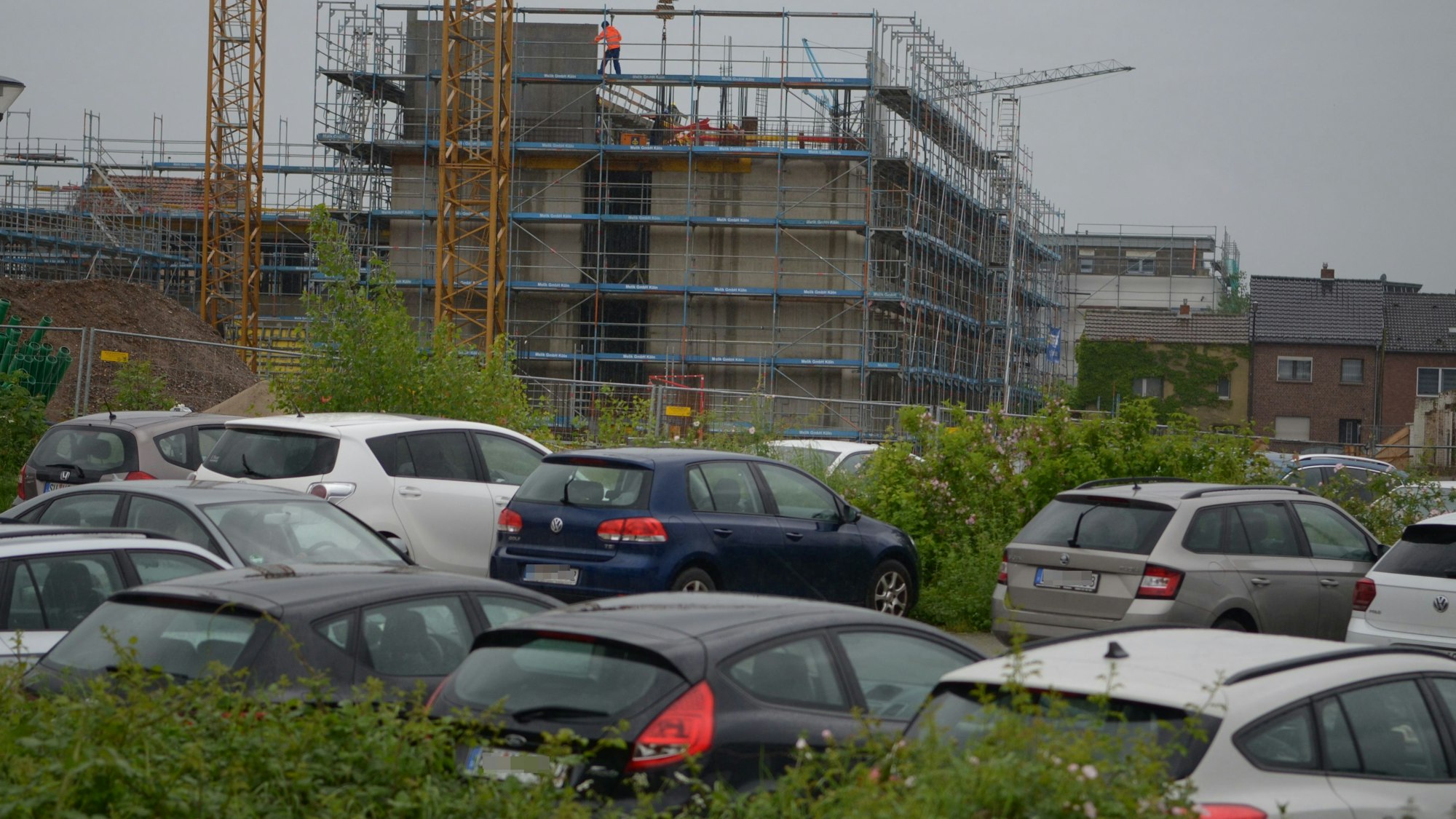 Im Vordergrund stehen in Reihen abgestellte Autos, dahinter die Baustelle mit dem eingerüsteten Rohbau.