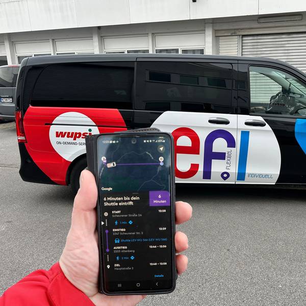 Ein „Efi“-Fahrzeug steht auf einem Parkplatz. Eine Hand hält davor ein Smartphone mit der „Efi“-Bestell-App.