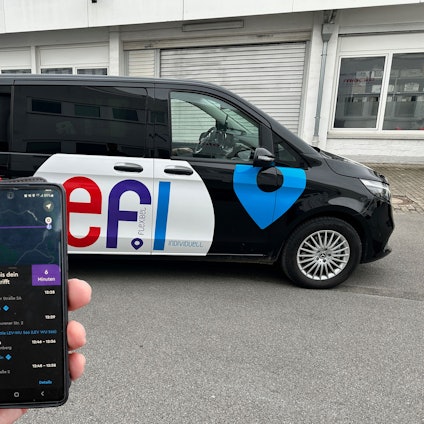 Ein „Efi“-Fahrzeug steht auf einem Parkplatz. Eine Hand hält davor ein Smartphone mit der „Efi“-Bestell-App.