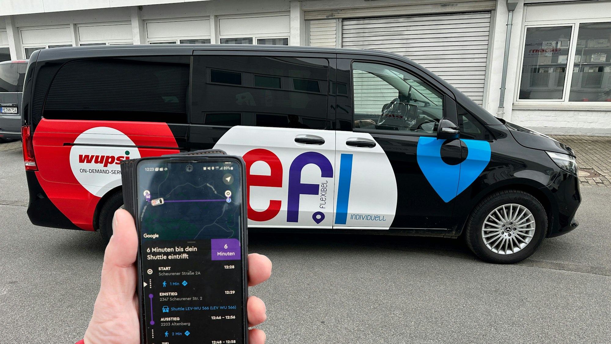 Eine Hand hält ein Handy, aus dem eine Fahrtstrecke aus einer App angezeigt wird. Dahinter steht ein Auto des Auf-Abruf-Verkehrsmittels „Efi“.