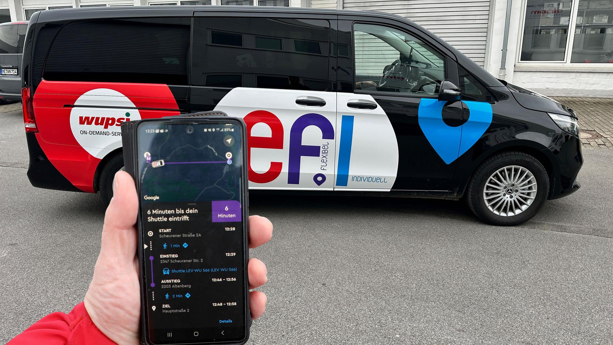 Ein „Efi“-Fahrzeug steht auf einem Parkplatz. Eine Hand hält davor ein Smartphone mit der „Efi“-Bestell-App.