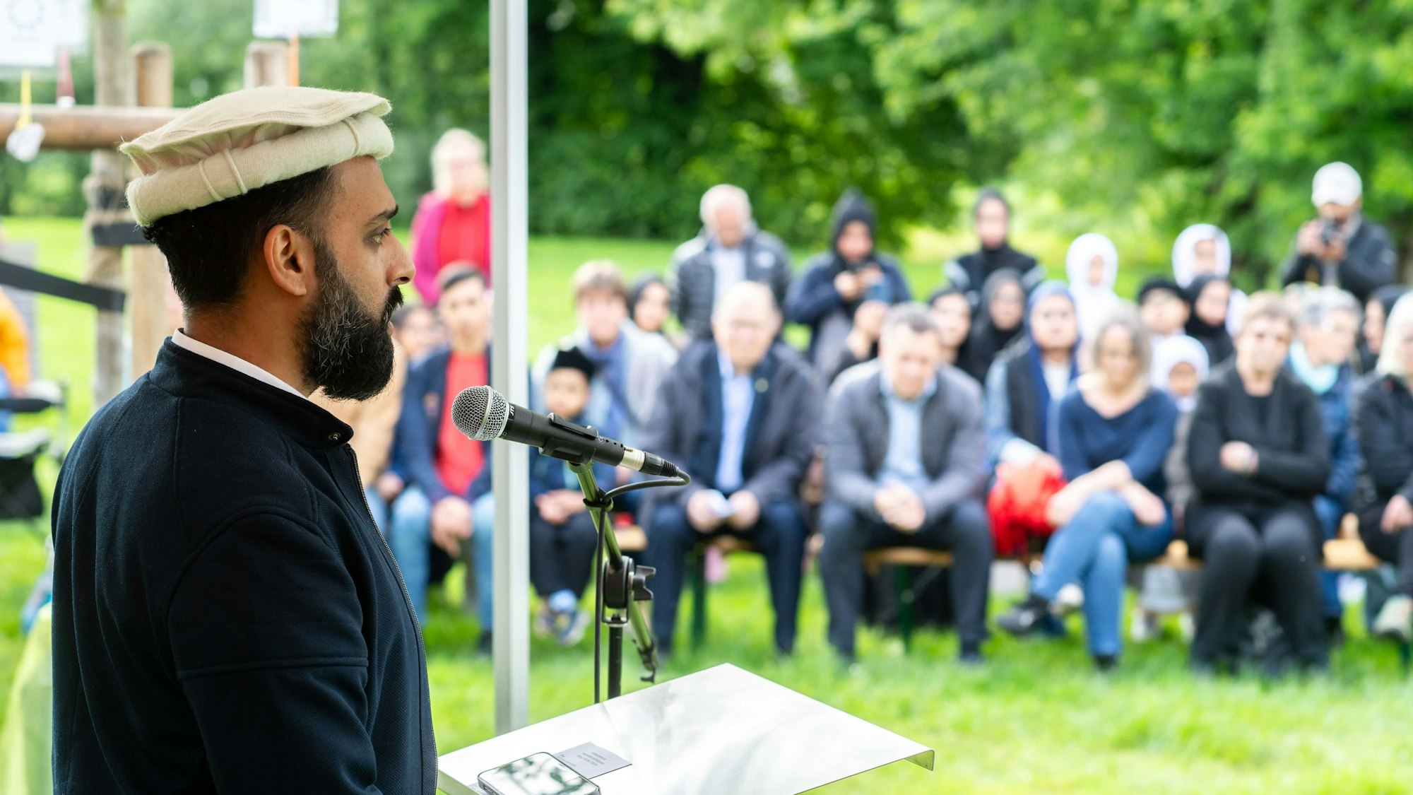 Der Redner spricht an einem Pult in ein Mikrofon, im Bildhintergrund sitzen Zuhörerinnen und Zuhörer.