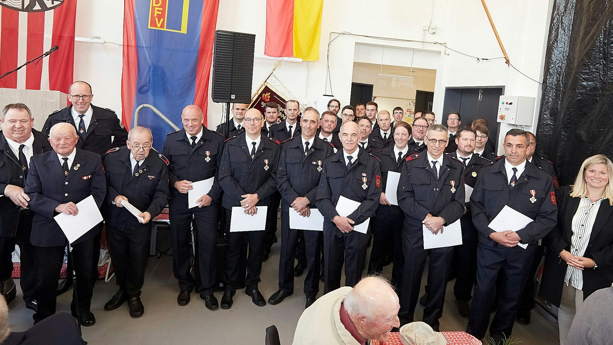 Zahlreiche Feuerwehrleute der Gemeinde Blankenheim stehen in Uniform in einer mit Fahnen geschmückten Halle. Sie halten Urkunden in den Händen. Rechts ist Bürgermeisterin Jennifer Meuren zu sehen.