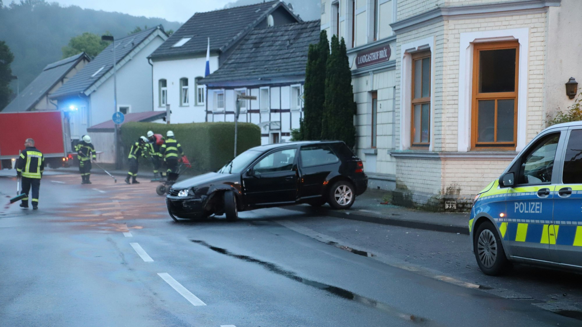 Ein Auto steht vor einem Haus, das linke Vorderrad steht quer. Feuerwehrleute verteilen Bindemittel auf der Ölspur.