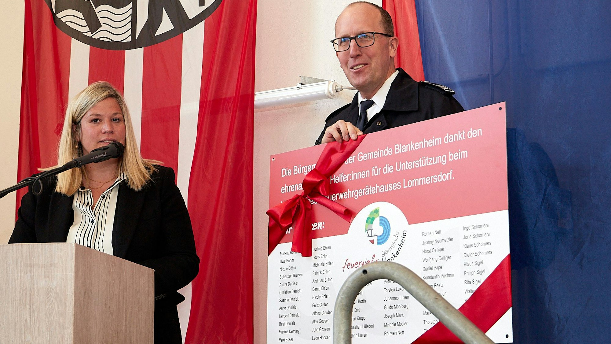 Blankenheims Bürgermeisterin Jennifer Meuren steht links an einem Pult mit einem Mikrofon, neben ihr steht der Lommersdorfer Löschgruppenführer, der eine Tafel in der Hand hält.