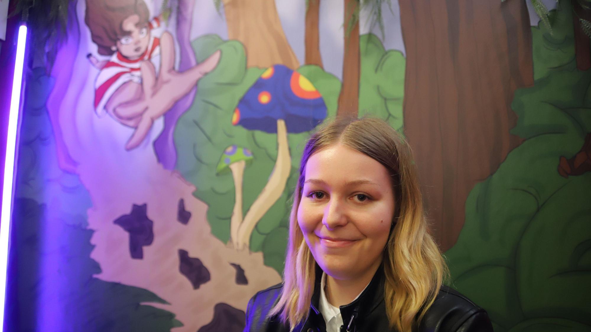 Kristina Hoffmann steht vor einer gemalten Szene aus ihrem Buch„ Amalia und die Reise durch Eldoria“.